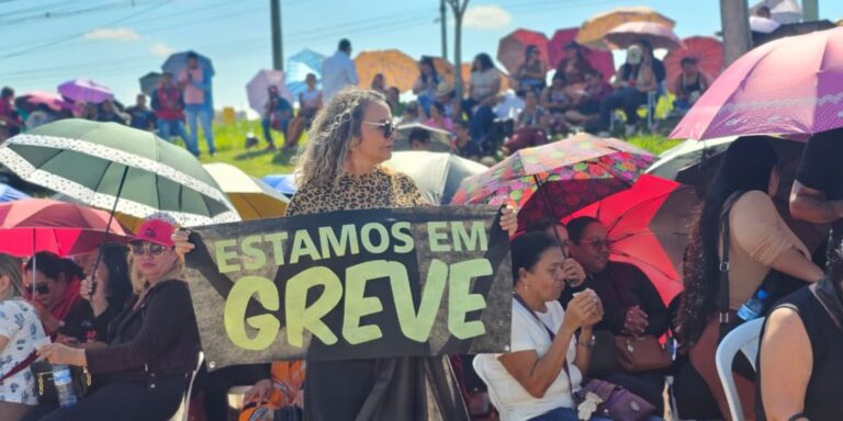 Mulher em primeiro plano segurando um cartaz no meio de uma multidão de manifestantes com uma faixa escrito estamos em greve ao fundo.