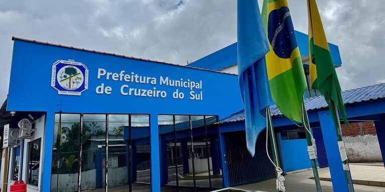 Fachada da Prefeitura Municipal de Cruzeiro do Sul sob o sol, com bandeiras hasteadas na entrada principal do prédio administrativo.